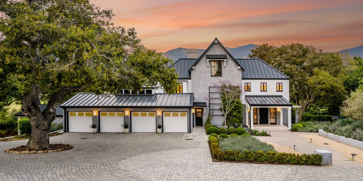 Modern luxury home with three-car garage and large tree in front at sunset.